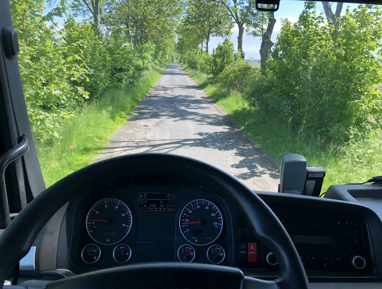 Widok z kabiny kierowcy ciężarówki na wiejską drogę otoczoną zielenią, z widoczną deską rozdzielczą i kierownicą.