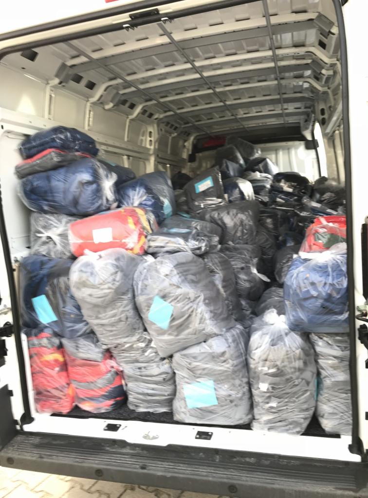 Wnętrze białego busa wypełnione po brzegi workami z odzieżą, niektóre worki z kolorowymi ubraniami, zabezpieczone folią, gotowe do transportu.
