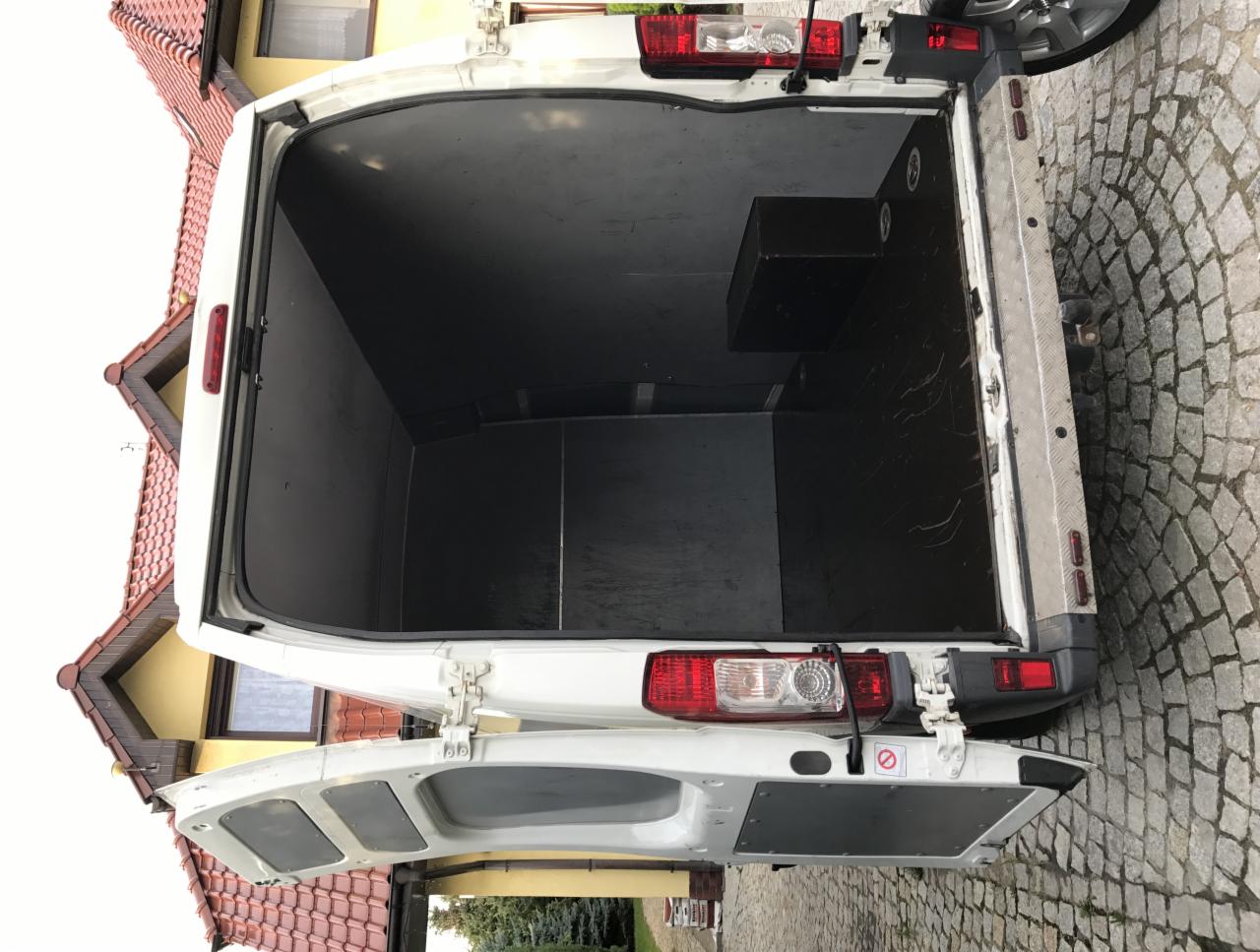 Wnętrze puste przestrzeni ładunkowej białego busa z otwartymi drzwiami, widok z tyłu na czarną podłogę i boczną skrzynkę, na zewnątrz kostka brukowa i fragment budynku.