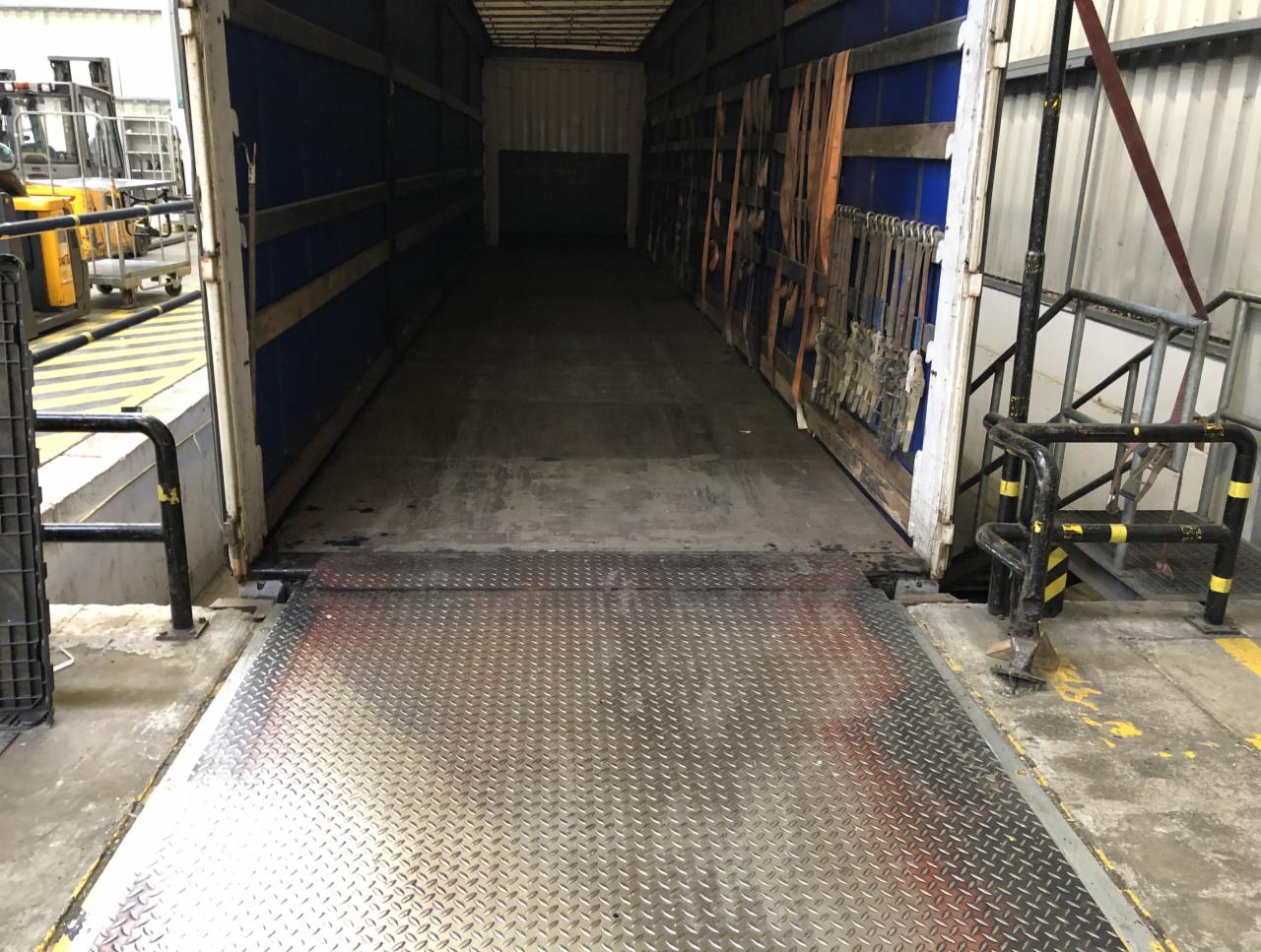 Wnętrze naczepy z rampą załadunkową, widoczne pasy mocujące i wózek widłowy w tle.