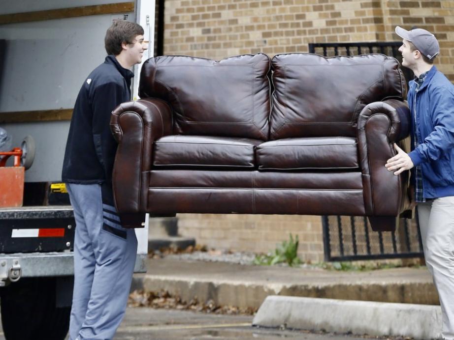 Dwóch mężczyzn wnosi skórzaną kanapę do ciężarówki przeprowadzkowej na tle ceglanego budynku i metalowego ogrodzenia. Widoczny fragment wnętrza pojazdu.