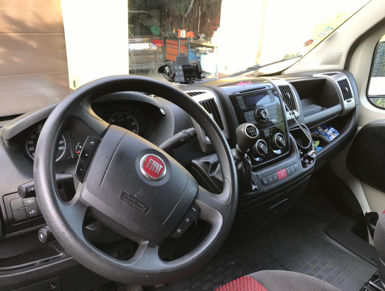 Wnętrze kabiny Fiata Ducato z widoczną kierownicą z logo, deską rozdzielczą, radiem i dźwignią zmiany biegów, uchwycone z perspektywy kierowcy.