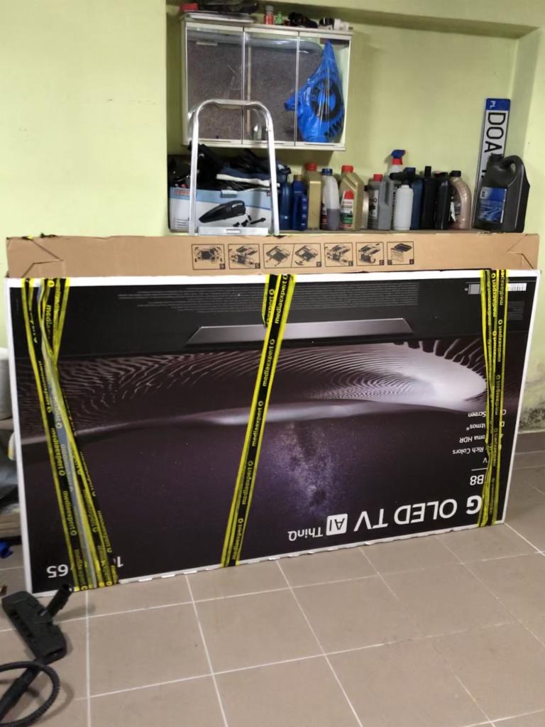 Karton z telewizorem OLED, zabezpieczony żółtymi taśmami, stoi w pomieszczeniu przypominającym garaż lub magazyn, z widocznymi narzędziami i pojemnikami na półkach w tle.