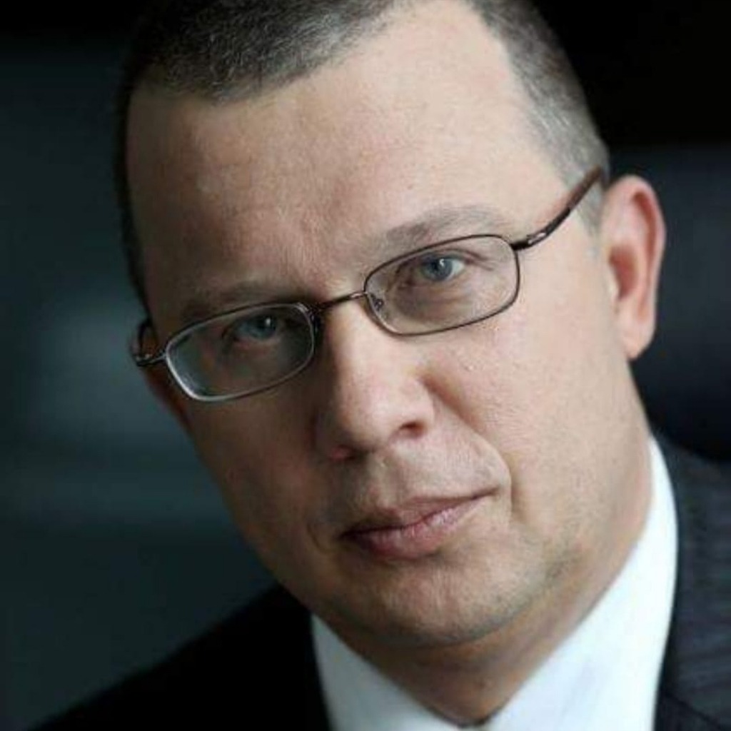 Portret mężczyzny w garniturze i okularach, skierowany lekko w prawo, z delikatnym uśmiechem.