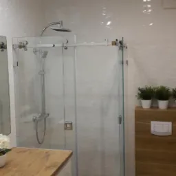 Nowoczesna łazienka z kabiną prysznicową typu walk-in, wykończona białymi płytkami z delikatnym wzorem i drewnianymi akcentami. Kabina z przesuwnymi drzwiami, deszczownica i odpływ liniowy w podłodze.