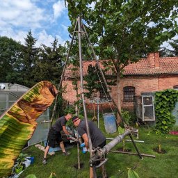 TBR SP Z O.O. - Dwóch mężczyzn pracuje przy metalowej konstrukcji w ogrodzie, prawdopodobnie podczas wiercenia; w tle szklarnia, budynek z czerwonej cegły i bujna roślinność.