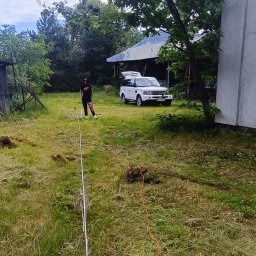 TBR SP Z O.O. - Pomiar działki przed rozpoczęciem prac ziemnych, widoczna rozciągnięta linka pomiarowa i wykopane dołki, w tle kobieta i biały samochód terenowy.