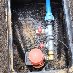 Instalacja pompy hydroforowej w studzience z zaworami kulowymi i niebieską rurą PE, widok z góry, czarna mata wewnątrz studzienki.