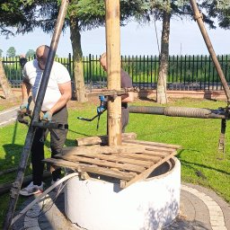TBR SP Z O.O. - Dwóch mężczyzn w trakcie montażu konstrukcji nad betonową studnią kręgową na trawniku, widoczne narzędzia i elementy konstrukcyjne, w tle ogrodzenie posesji i drzewa.