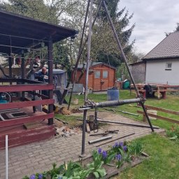 TBR SP Z O.O. - Metalowa wiertnica trójnożna w ogrodzie, z rurami na brukowanym podłożu, obok kwitnących fioletowych hiacyntów. W tle drewniana altana i pracownik.