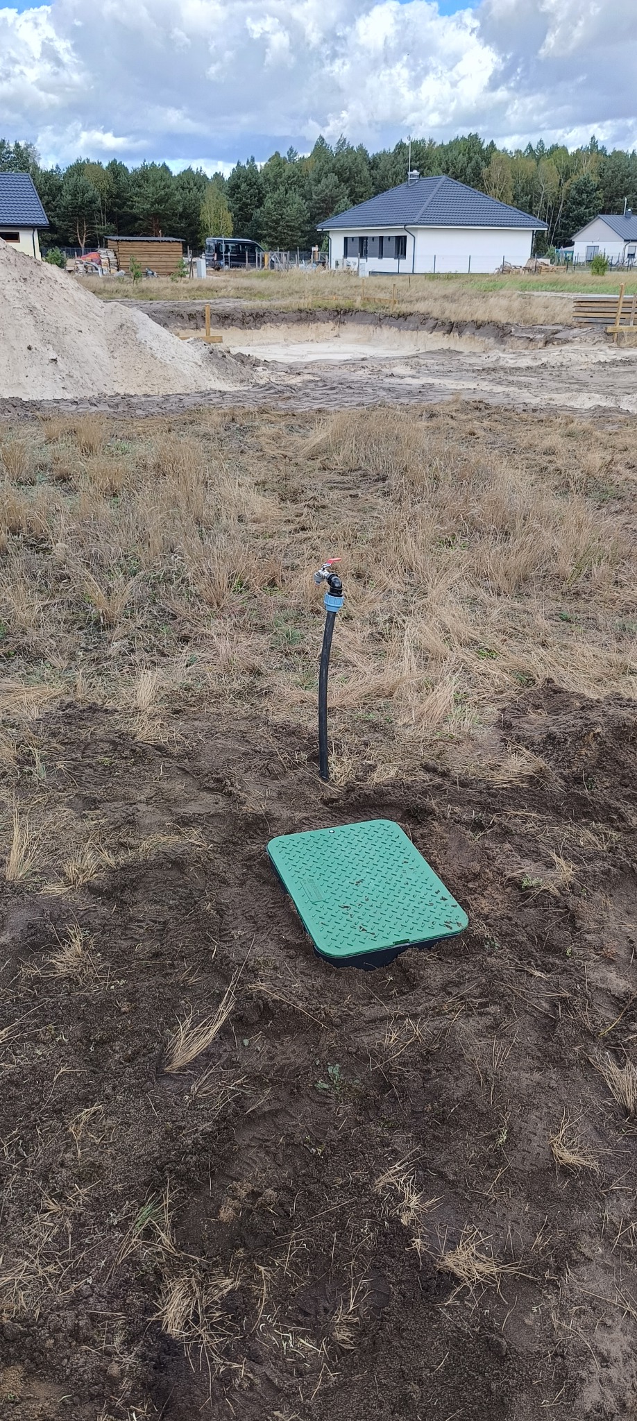 Zielona pokrywa studzienki rewizyjnej z kranem wystającym z ziemi na tle placu budowy z nowymi domami i stertą piasku.