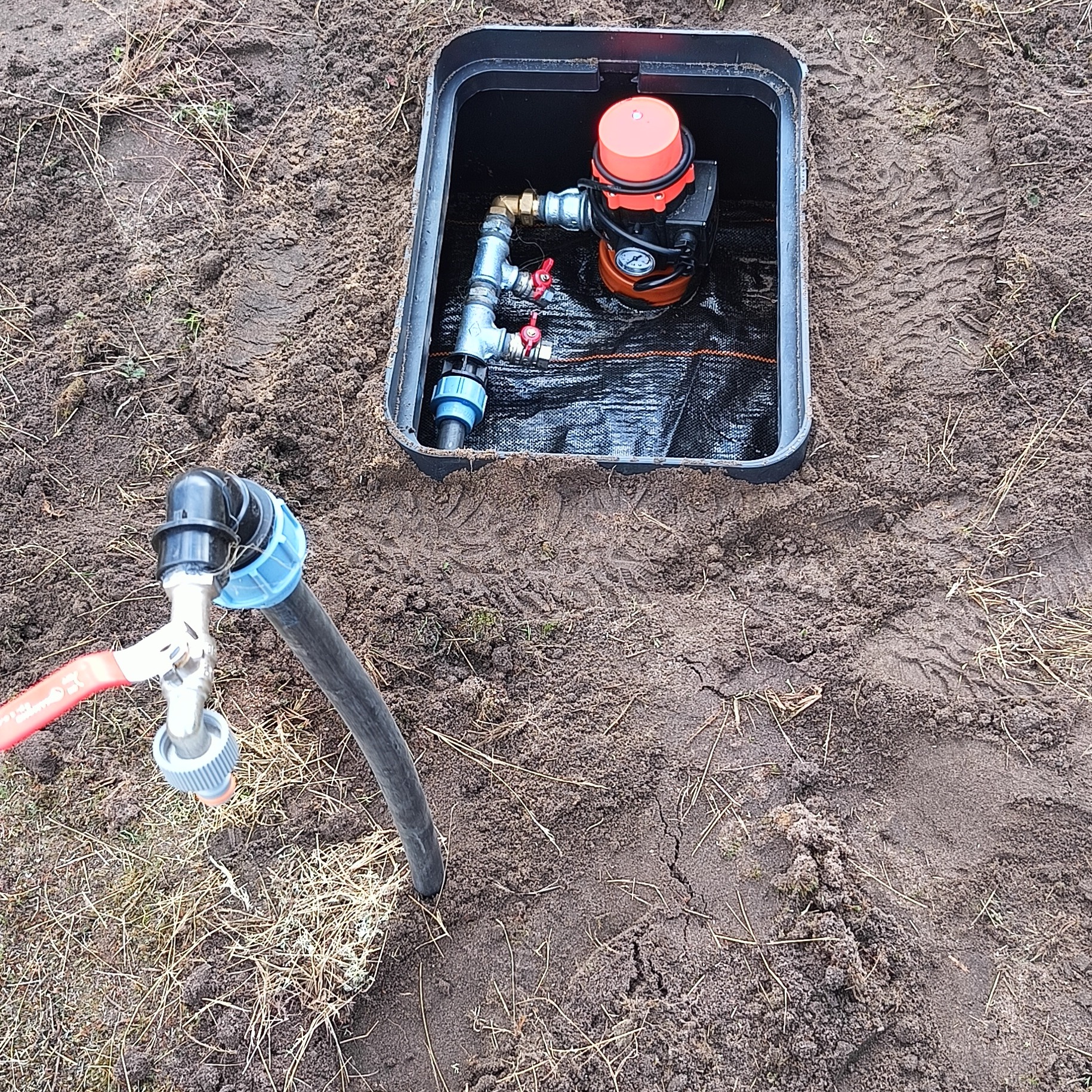 Instalacja pompy hydroforowej w czarnej studzience na ziemi, z kranem i wężem doprowadzającym wodę, widoczne ślady opon w ziemi.