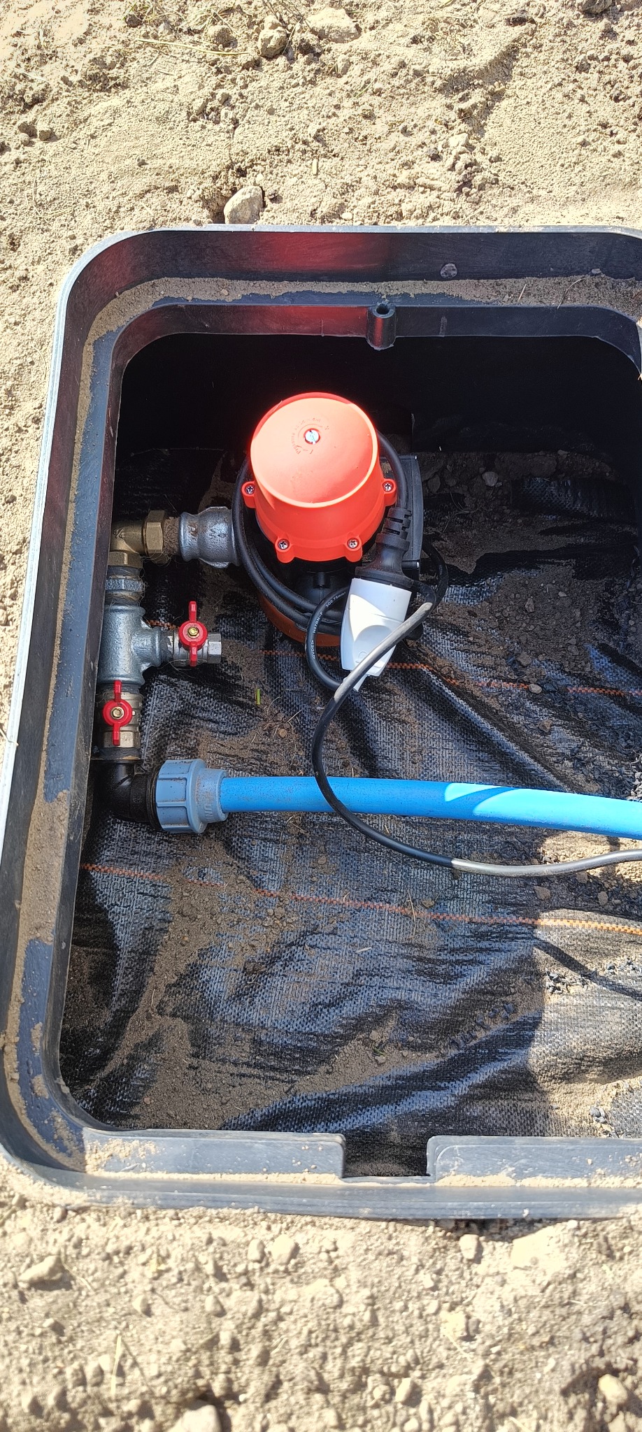 Pompa głębinowa z czerwonym korpusem i zaworami kulowymi w czarnej skrzynce inspekcyjnej, z niebieską rurą odprowadzającą wodę, zainstalowana na czarnej geowłókninie.