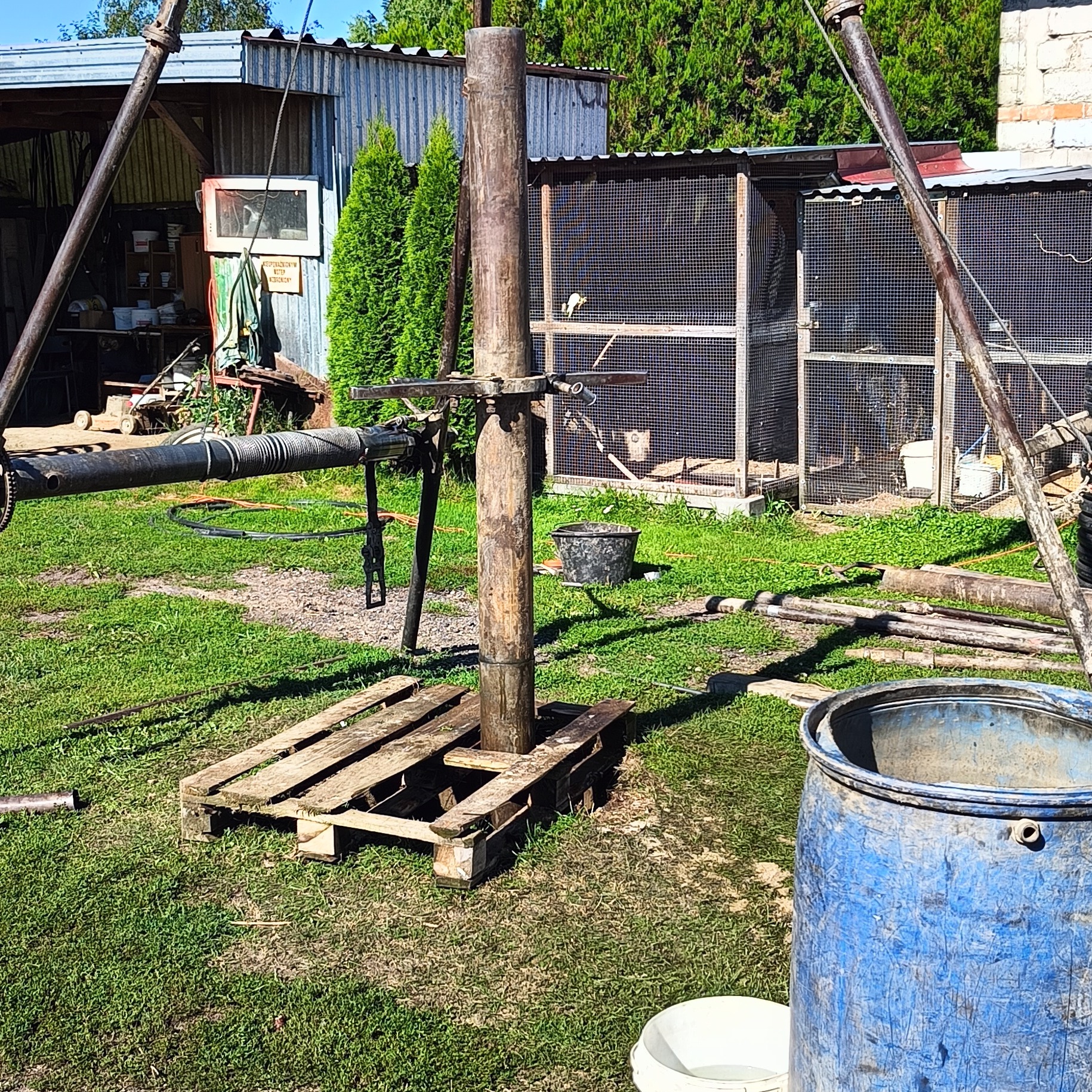 Sprzęt do wiercenia studni na trawiastym terenie, z widocznym fragmentem klatki dla ptaków w tle i niebieską beczką z przodu.