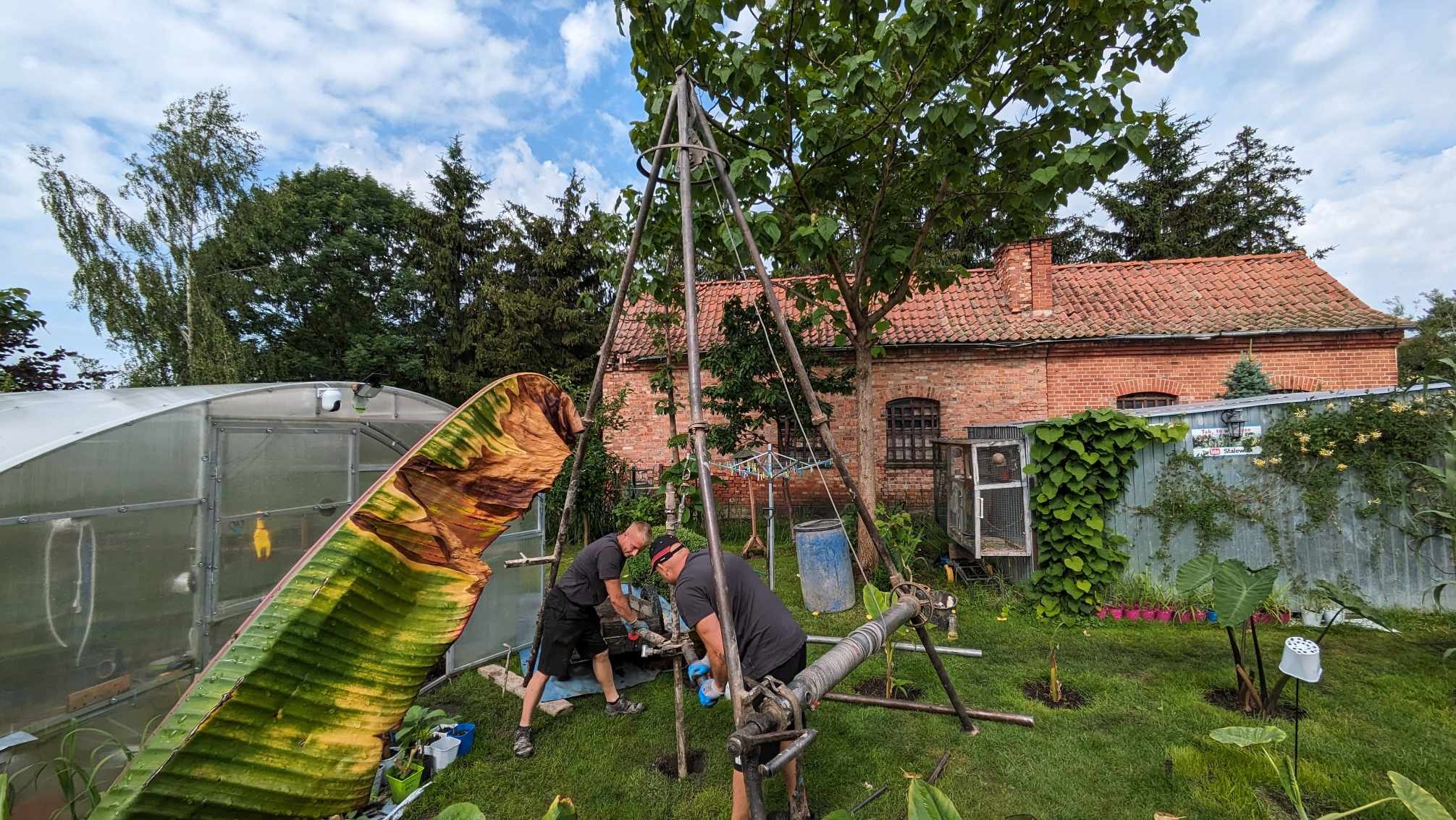 Dwóch mężczyzn pracuje przy metalowej konstrukcji w ogrodzie, prawdopodobnie podczas wiercenia; w tle szklarnia, budynek z czerwonej cegły i bujna roślinność.