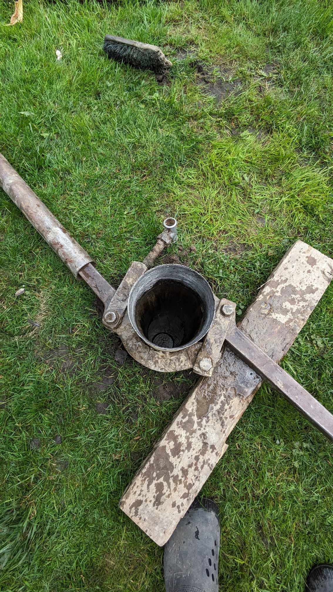 Metalowa rura z mechanizmem wiertniczym leżąca na trawie, obok widoczny fragment buta roboczego i szczotka.