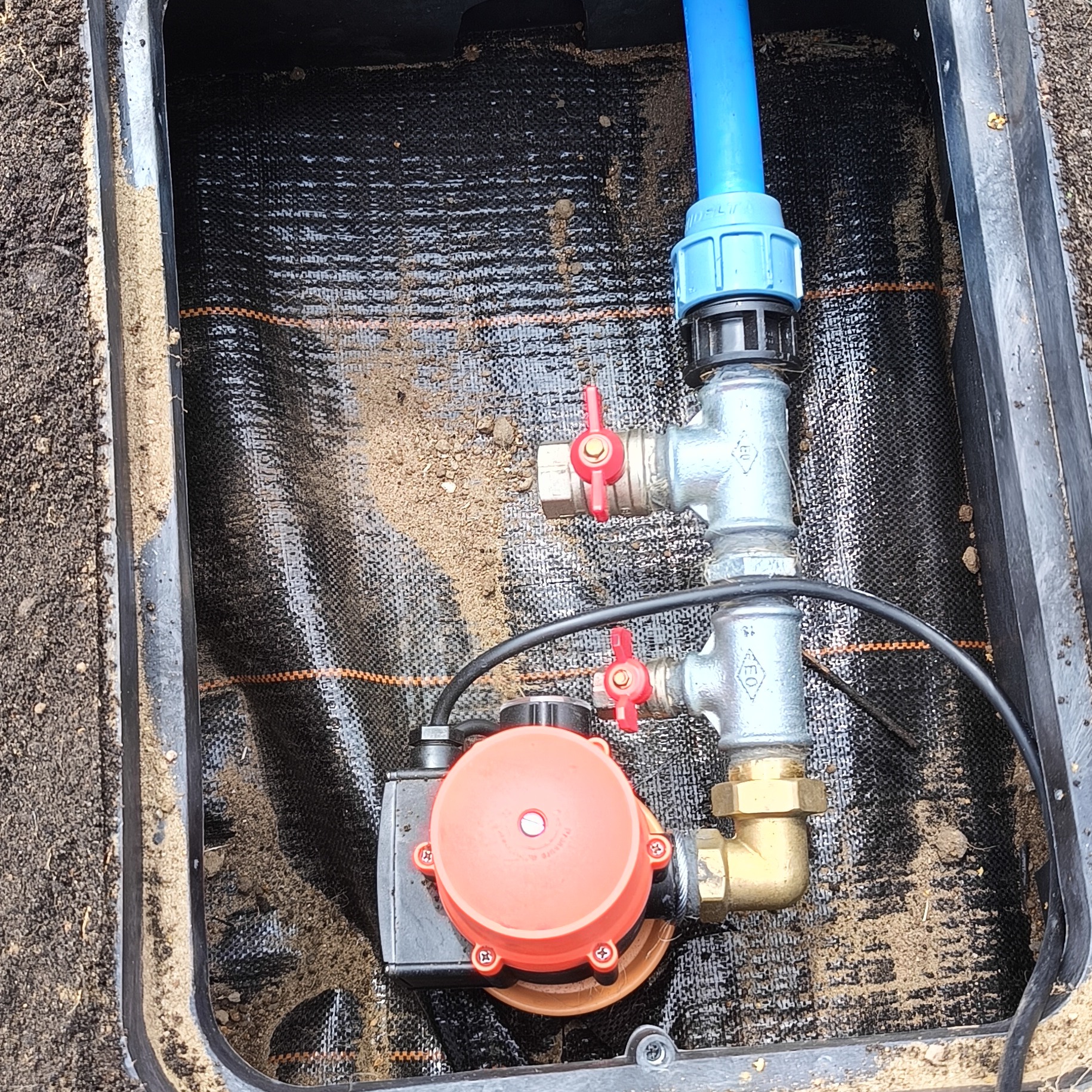 Instalacja pompy hydroforowej w studzience z zaworami kulowymi i niebieską rurą PE, widok z góry, czarna mata wewnątrz studzienki.