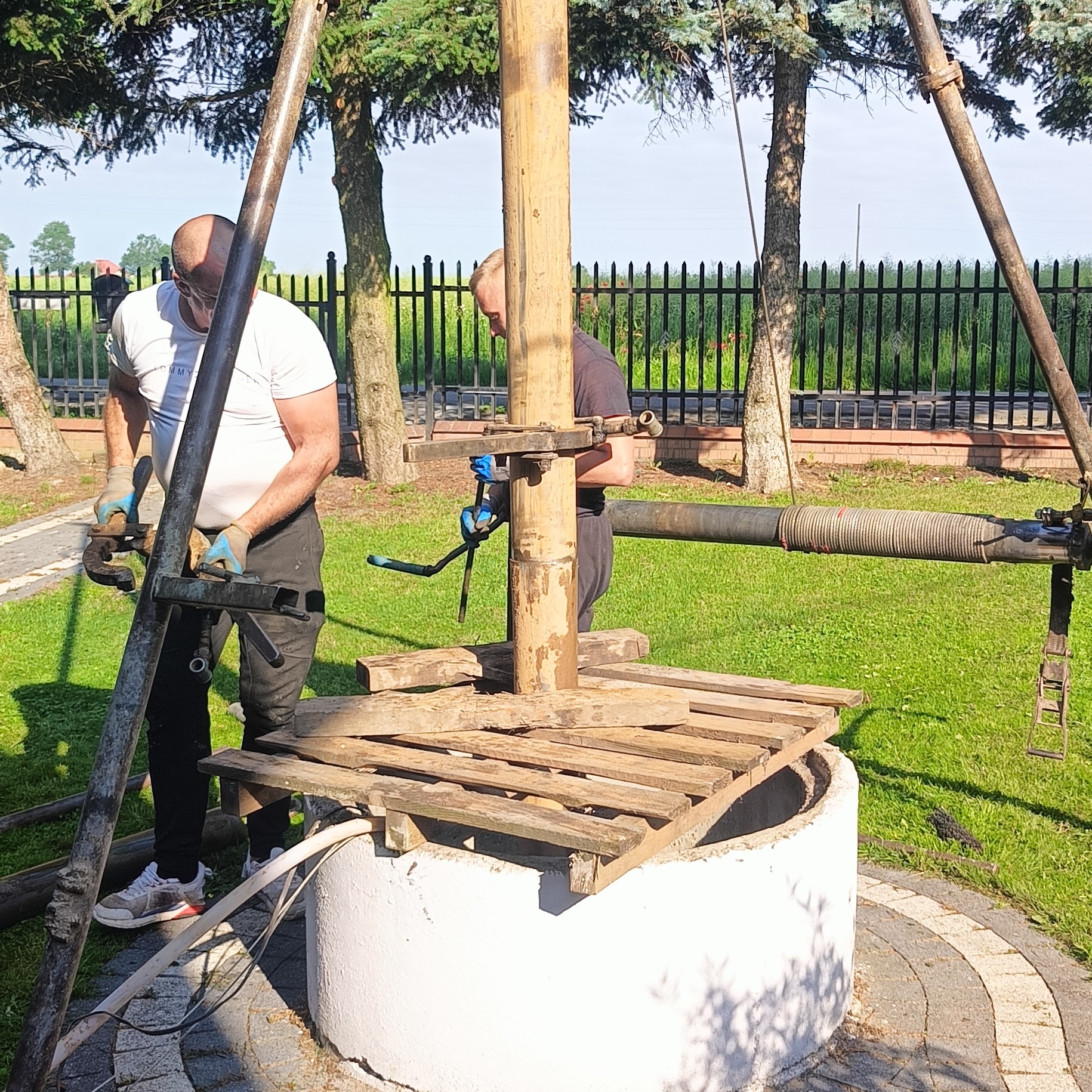 Dwóch mężczyzn w trakcie montażu konstrukcji nad betonową studnią kręgową na trawniku, widoczne narzędzia i elementy konstrukcyjne, w tle ogrodzenie posesji i drzewa.