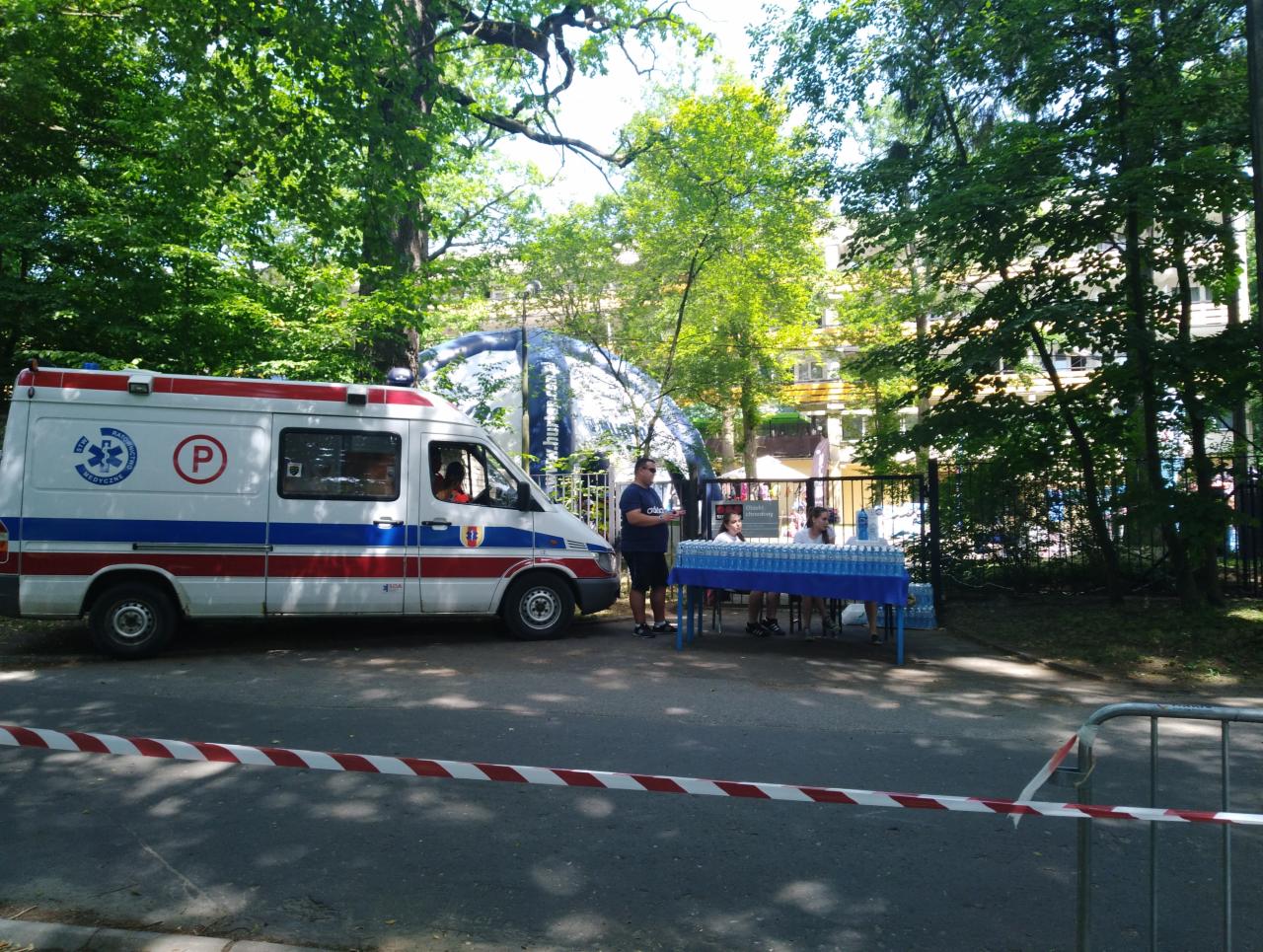 Ambulans z symbolem 'P' na tle parku, obok stolik z butelkami wody, prawdopodobnie punkt medyczny podczas wydarzenia sportowego.