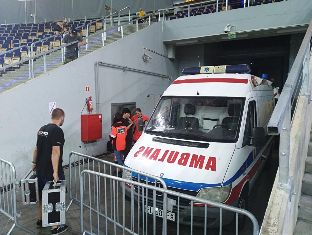 Ambulans zaparkowany w tunelu stadionu, personel medyczny w pomarańczowych kamizelkach obok pojazdu, mężczyzna przenosi sprzęt w metalowych skrzynkach.