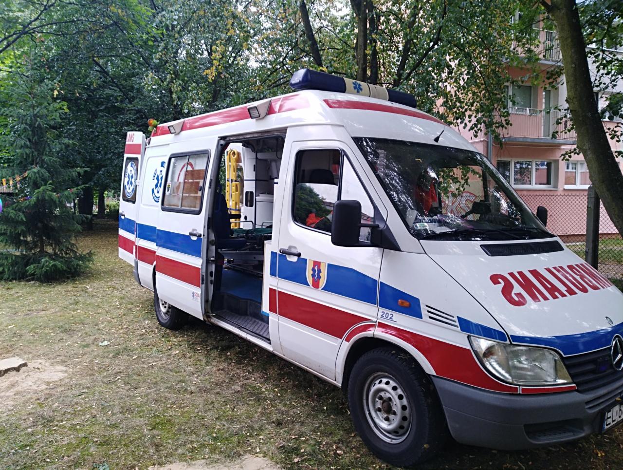 Ambulans z otwartymi drzwiami bocznymi, widoczny wózek transportowy wewnątrz, zaparkowany na trawie przed budynkiem.