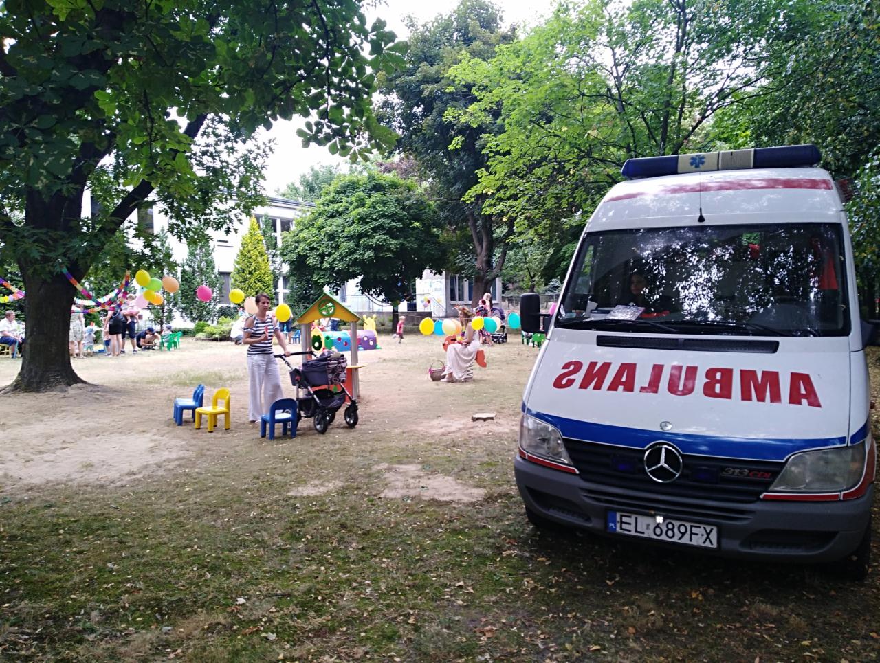 Ambulans zaparkowany na trawniku w otoczeniu bawiących się dzieci, z odwróconym napisem 'AMBULANS' na przedniej szybie.