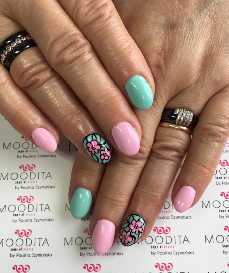 Dłonie z pastelowym manicure: różowe i miętowe paznokcie, na dwóch z nich czarne kontury kwiatów na miętowym tle, pierścionki na palcach, tło z logo salonu.