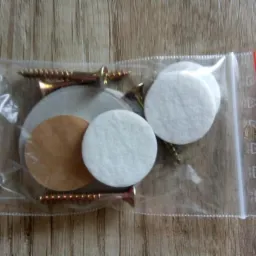 Foliowa torebka strunowa z zestawem akcesoriów: wkręty z płaskim łbem, białe podkładki filcowe i brązowe podkładki samoprzylepne na drewnianym tle.