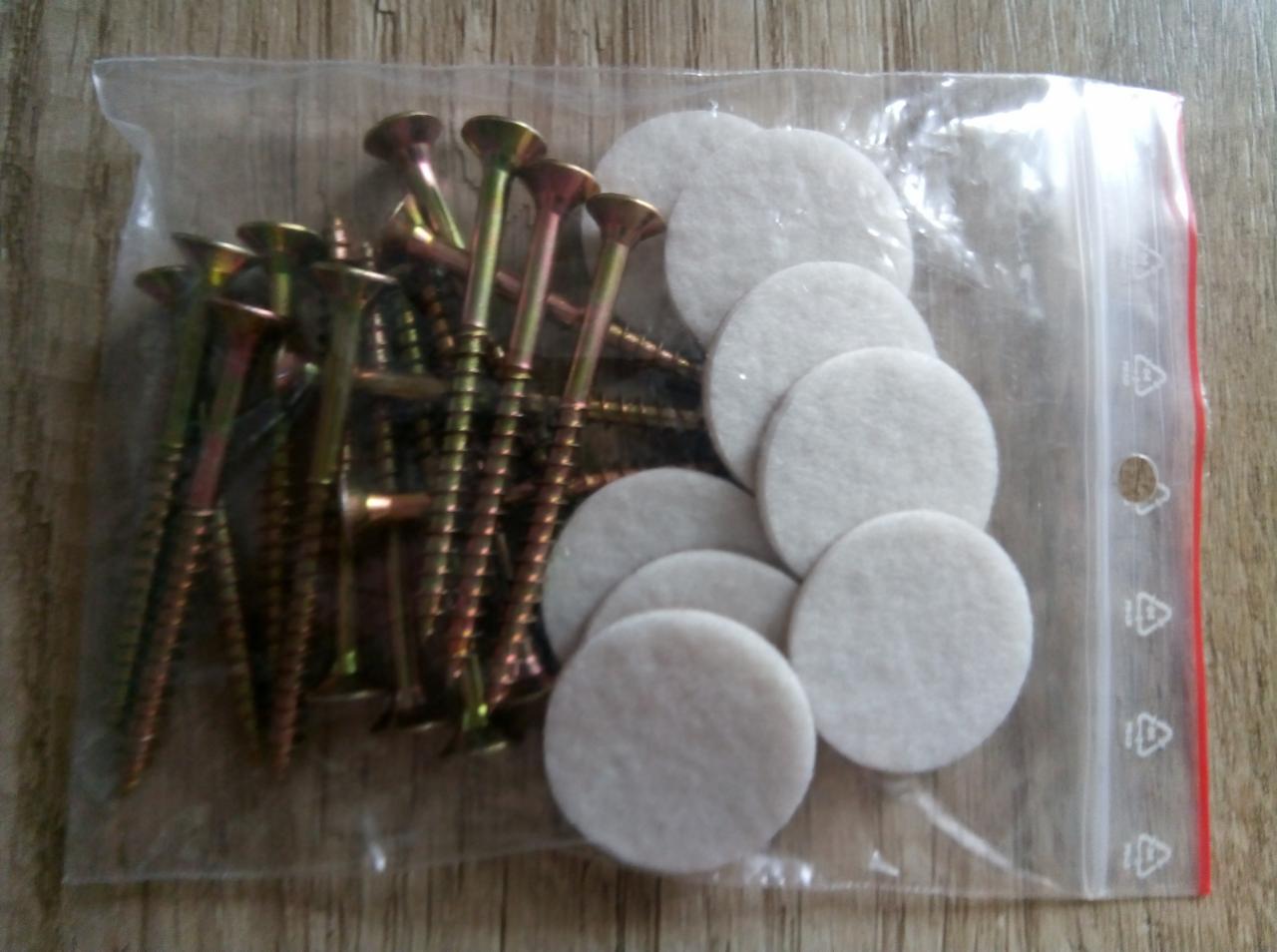 Zestaw montażowy: śruby z żółtego metalu i okrągłe, samoprzylepne podkładki filcowe w przezroczystym woreczku strunowym na drewnianym tle.