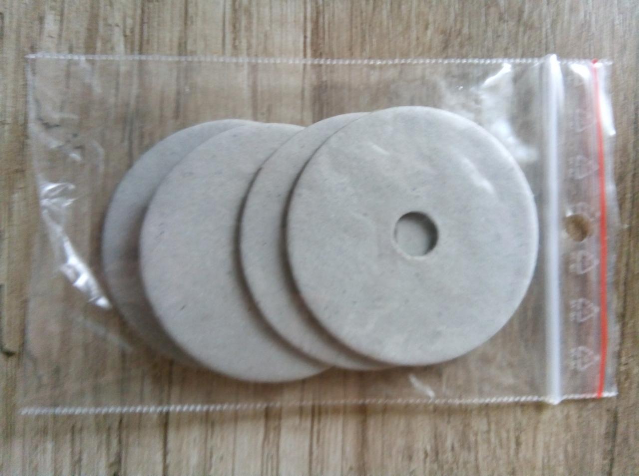 Szare, okrągłe podkładki z otworem w środku, ułożone jedna na drugiej w przezroczystym, plastikowym woreczku strunowym, widoczne na tle drewnianej powierzchni.