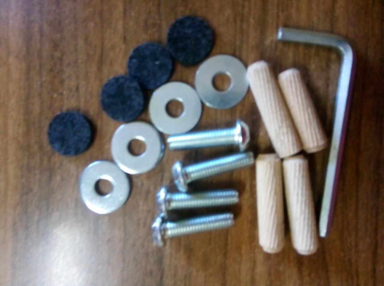 Zestaw akcesoriów montażowych: cztery śruby z okrągłymi łbami, cztery podkładki, cztery drewniane kołki, cztery czarne filcowe podkładki oraz klucz imbusowy, ułożone na drewnianej powierzchni.