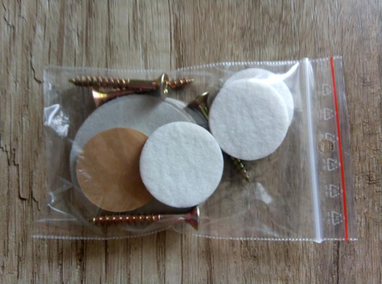 Foliowa torebka strunowa z zestawem akcesoriów: wkręty z płaskim łbem, białe podkładki filcowe i brązowe podkładki samoprzylepne na drewnianym tle.