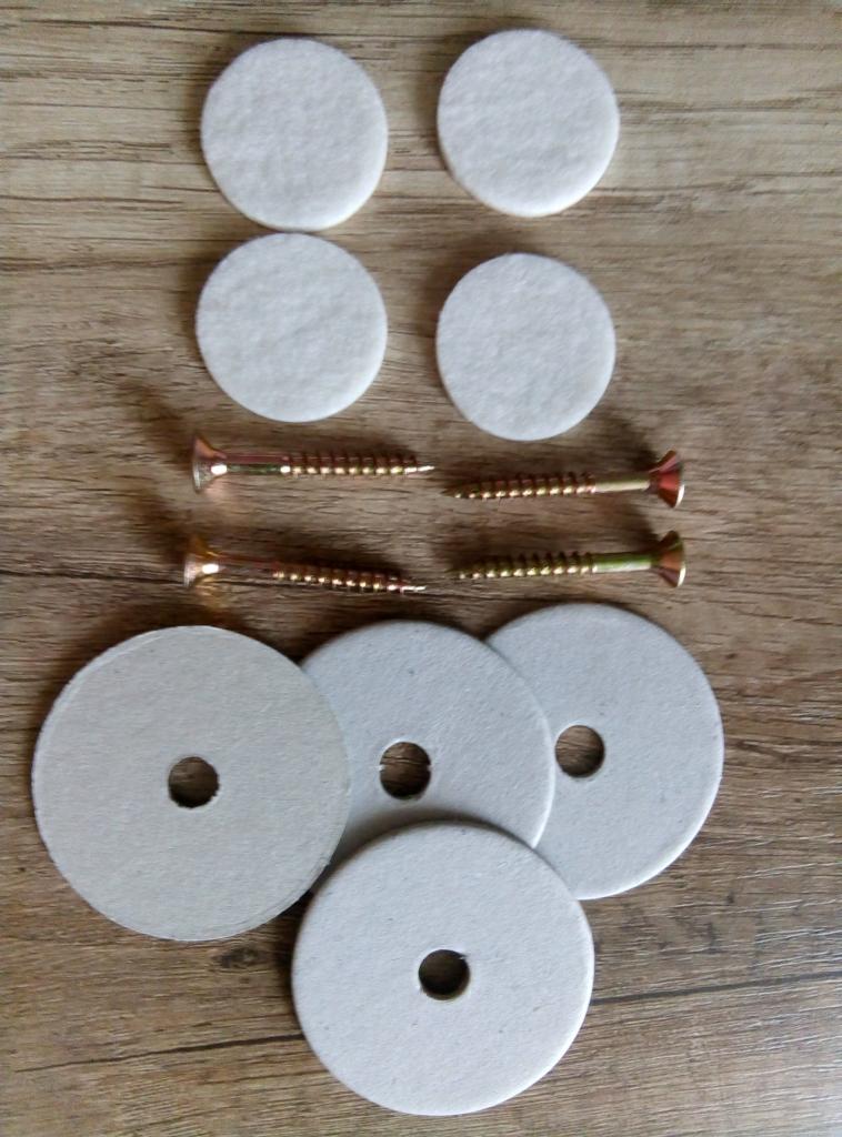 Zestaw akcesoriów do montażu: cztery białe filcowe podkładki, cztery podkładki z otworem oraz cztery śruby na drewnianym tle.