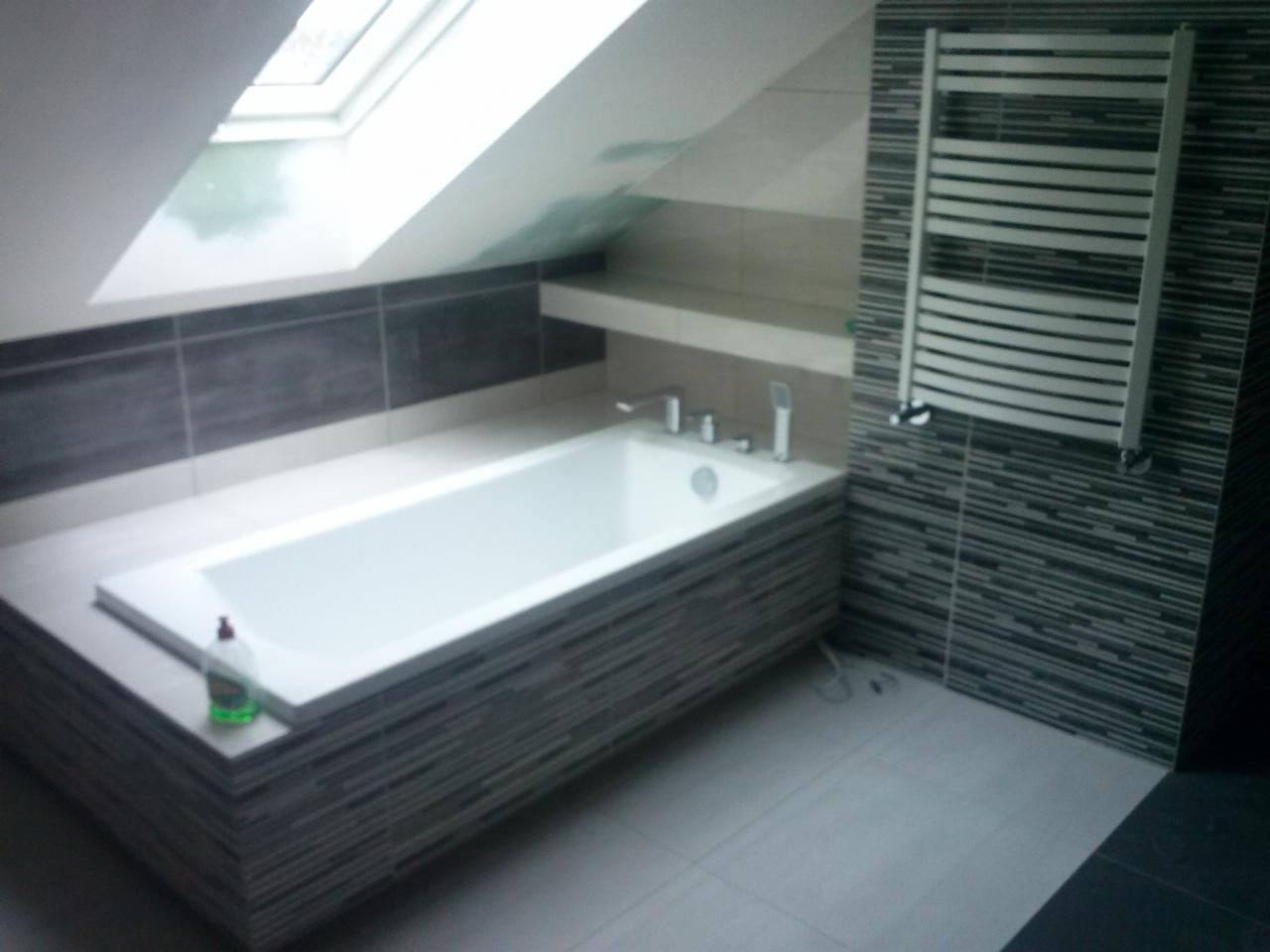 Biała wanna w zabudowie z mozaiki w łazience na poddaszu ze świetlikiem, obok grzejnik drabinkowy na ścianie z mozaiki.