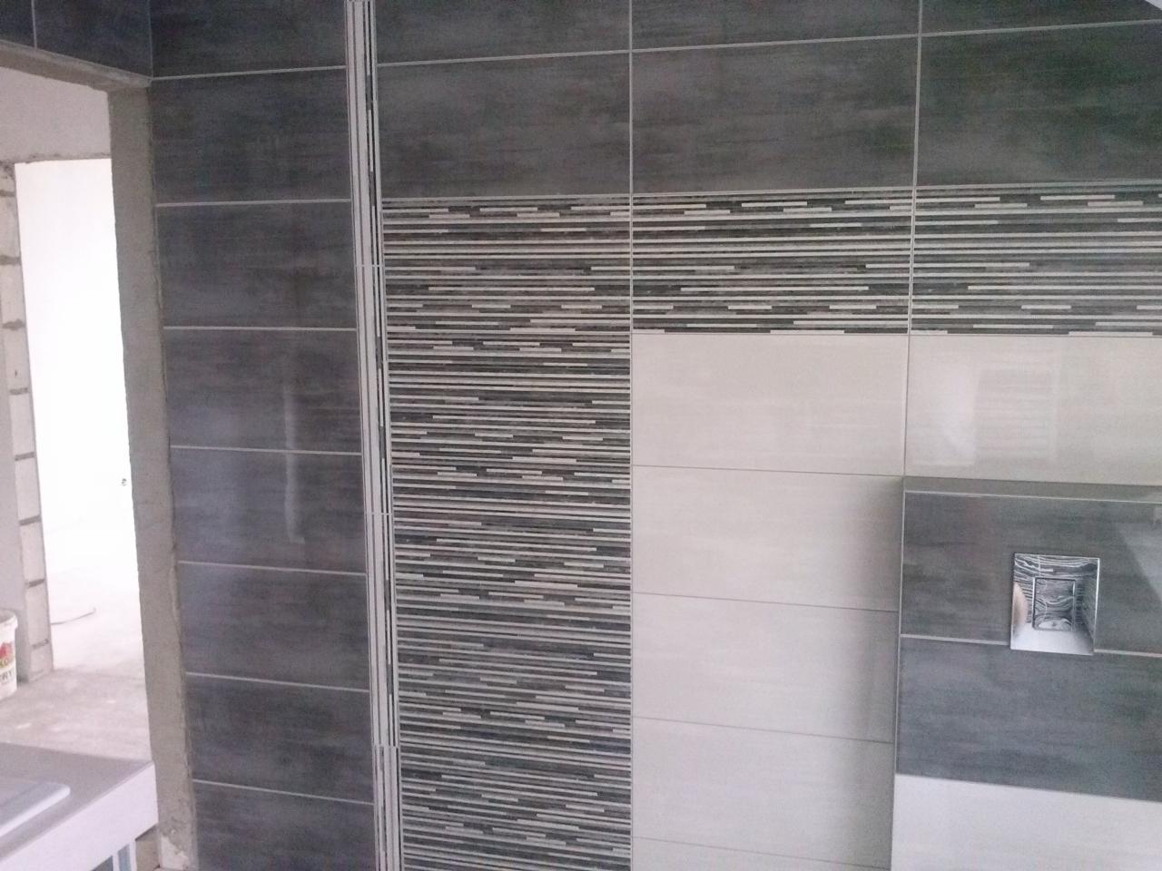 Ściana łazienki w trakcie wykańczania z szarymi i białymi płytkami, widoczny fragment stelaża podtynkowego WC, surowy stan wykończenia w tle.