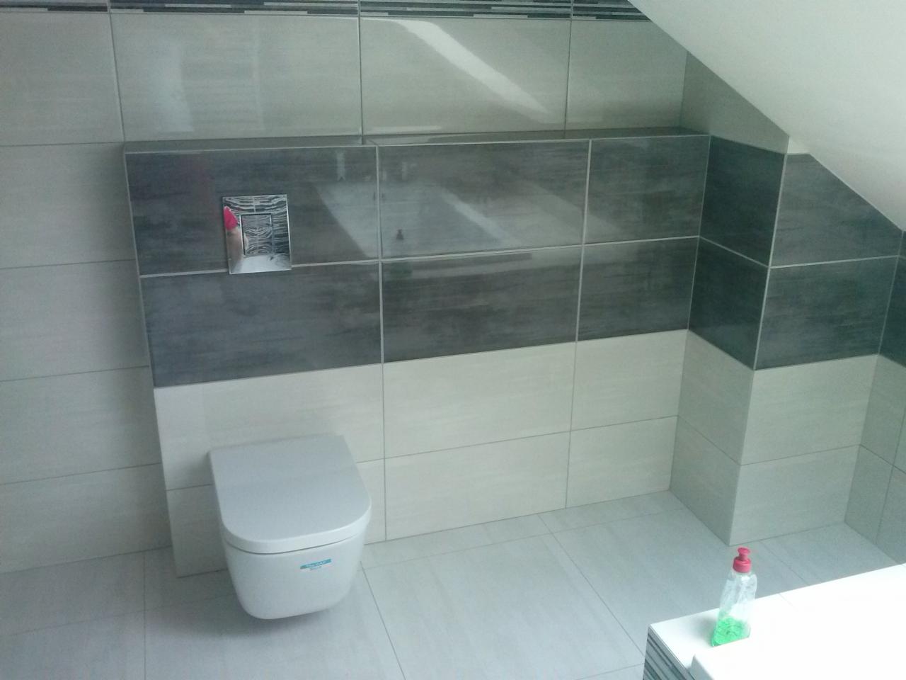 Podwieszana toaleta w łazience wykończonej płytkami w odcieniach szarości i bieli, widoczny fragment skosu dachu i butelka środka czyszczącego na blacie.