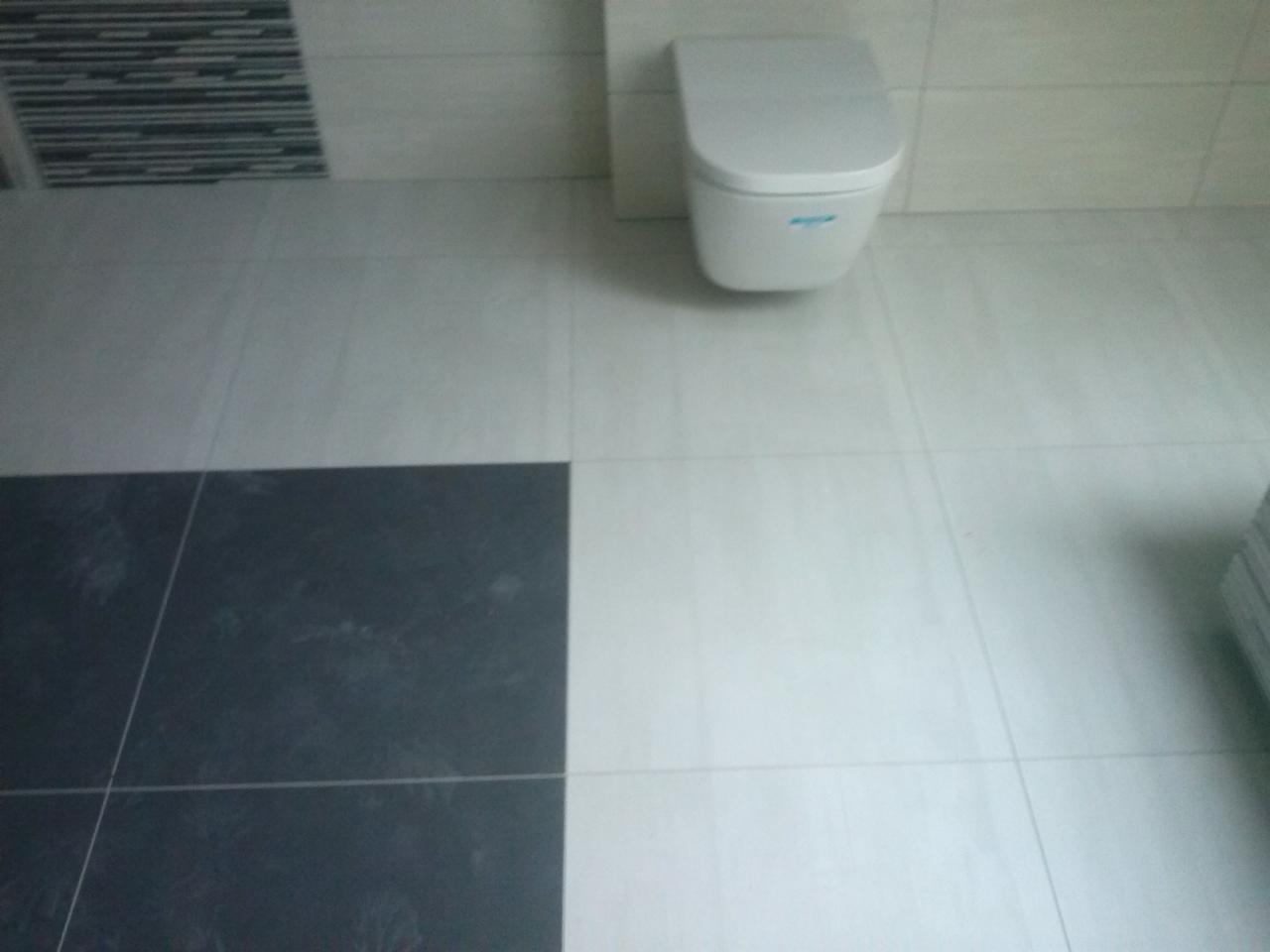 Wisząca toaleta z białą ceramiką na tle jasnych płytek ściennych i podłogowych, fragment podłogi z ciemnymi płytkami w lewym dolnym rogu.