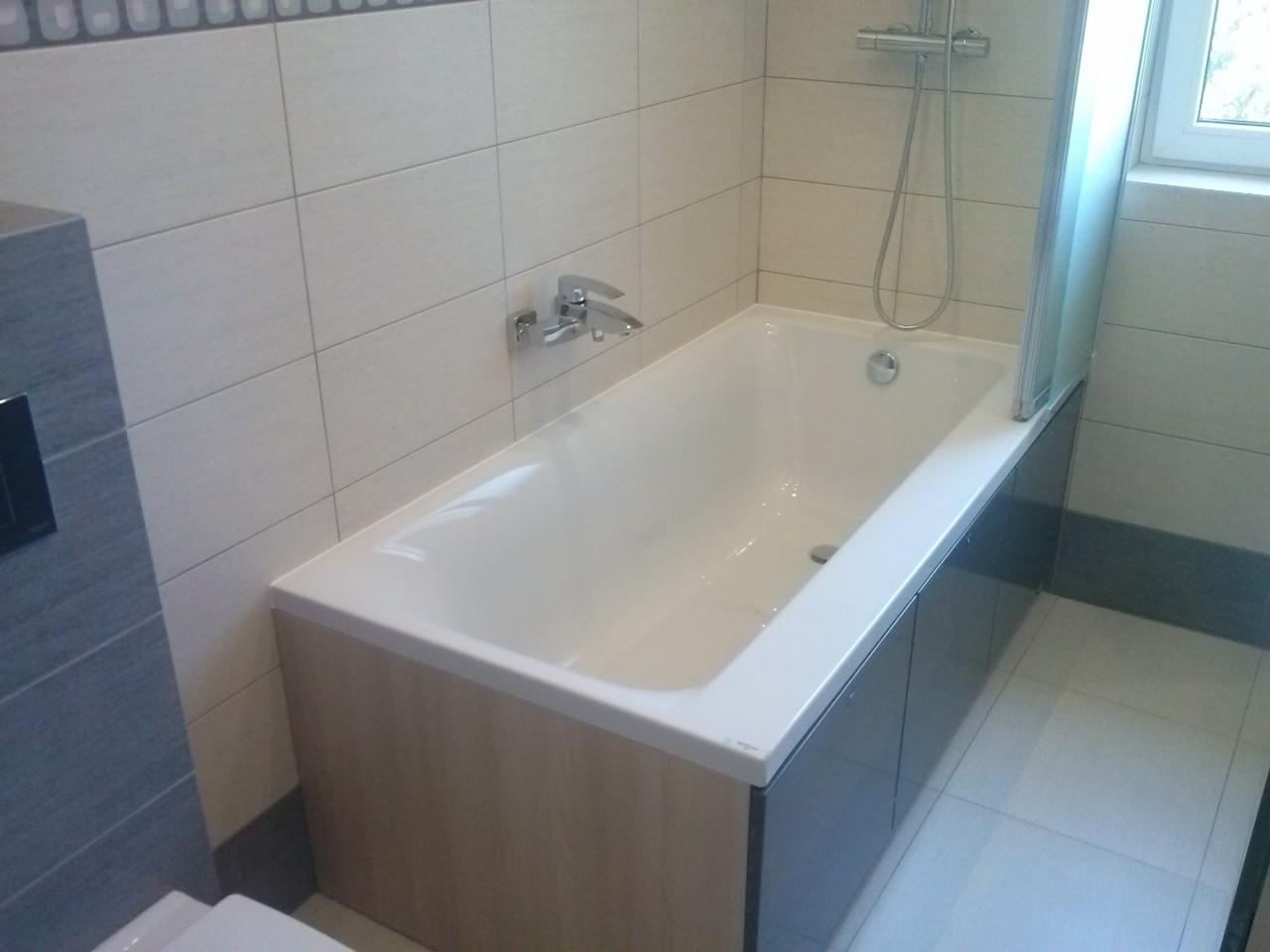 Biała wanna z zabudową w odcieniach szarości i beżu, chromowana bateria i słuchawka prysznicowa, jasne płytki na ścianach i podłodze, minimalistyczne wykończenie łazienki.