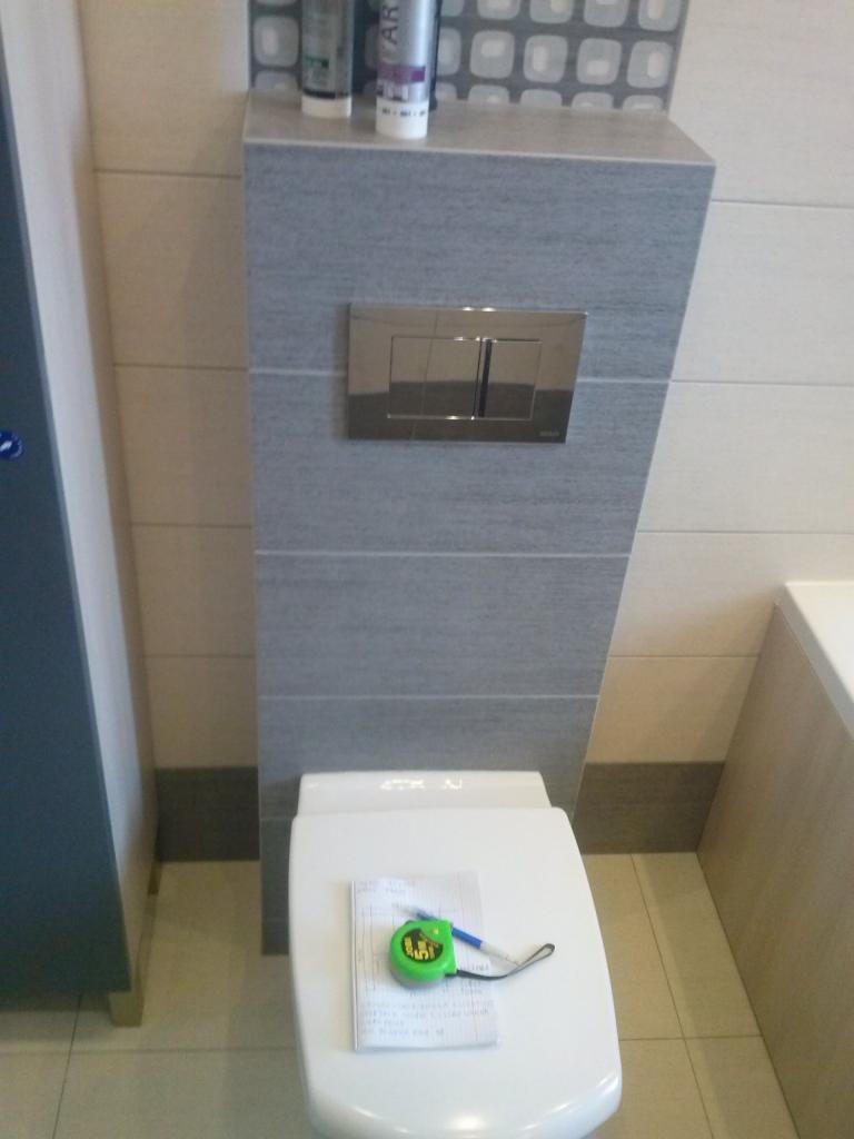 Kompakt WC z ukrytą spłuczką w zabudowie z szarych płytek, na desce leży notatnik z zieloną miarką zwijaną i długopisem.