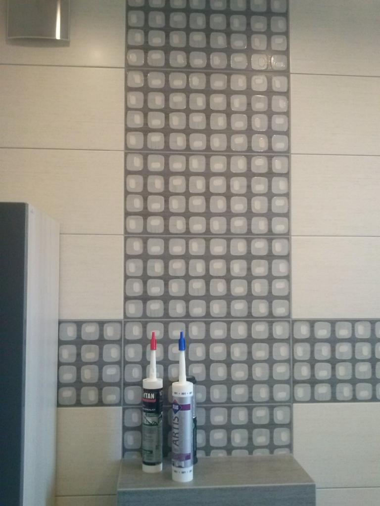 Ściana wyłożona jasnymi i szarymi płytkami ceramicznymi z dekoracyjnym pasem w kwadraty o zaokrąglonych rogach; dwie tuby z silikonem (czerwona i niebieska) stoją na półce.