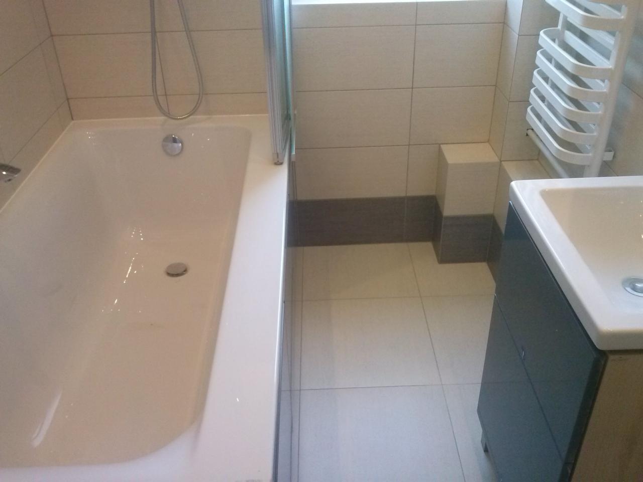 Wykończona łazienka z białą wanną, szklanym parawanem, umywalką z szafką w odcieniach szarości i grzejnikiem drabinkowym. Płytki w jasnych odcieniach beżu i szarości.