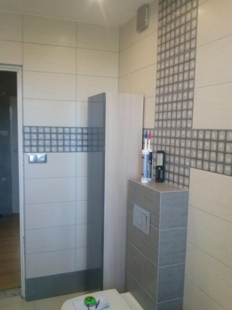 Wykończona łazienka z beżowymi i szarymi płytkami, dekoracyjnym pasem z motywem geometrycznym, zabudowaną spłuczką WC i akcesoriami na blacie. Na sedesie leżą notatki i miarka.