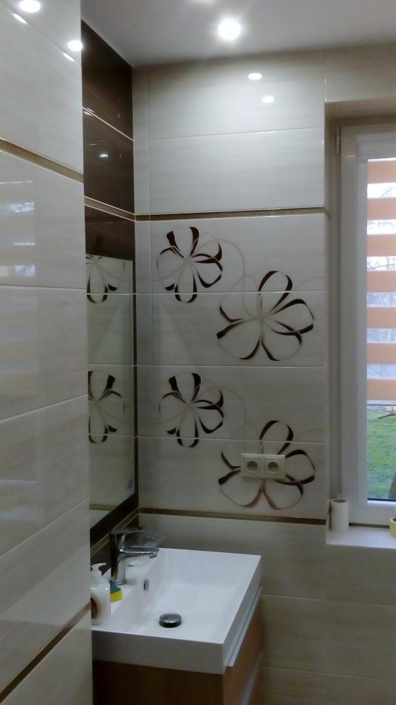 Jasna łazienka z umywalką o prostokątnym kształcie, baterią chromowaną i lustrem z brązowymi akcentami, ściany wyłożone płytkami z motywem kwiatowym oraz gniazdkiem elektrycznym.