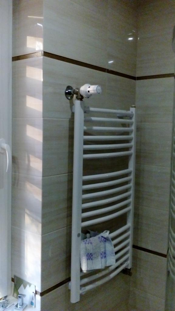 Biały, drabinkowy grzejnik łazienkowy z termostatem, zamontowany na ścianie wyłożonej beżowymi płytkami z ciemnobrązowym paskiem dekoracyjnym; na grzejniku zawieszony ręcznik w kratę.