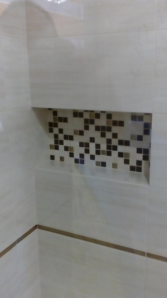 Wykończona wnęka w ścianie łazienki, wyłożona beżowymi płytkami z dekoracyjną mozaiką z brązowych i beżowych kwadratów wewnątrz, oraz cienkim brązowym paskiem dekoracyjnym w dolnej części ściany.