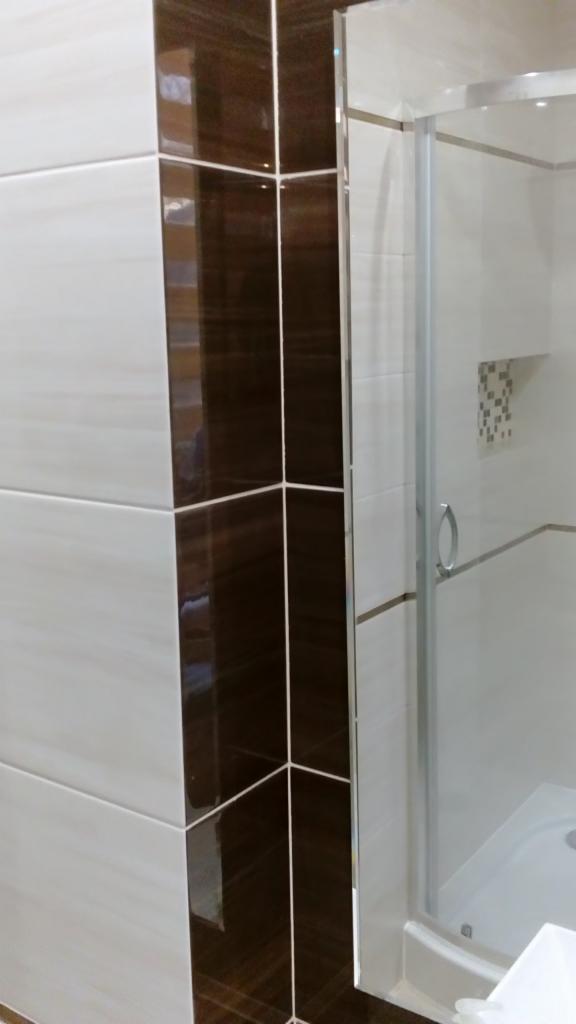 Szczegółowe ujęcie narożnika kabiny prysznicowej z białymi i brązowymi płytkami, widoczne srebrne wykończenia i uchwyt kabiny.