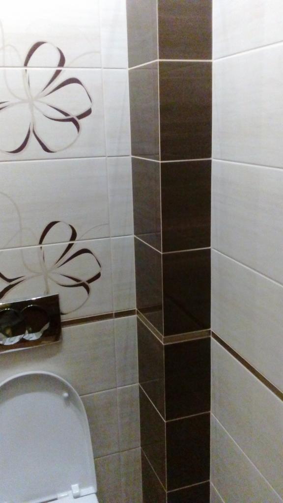 Wykończona łazienka z brązowo-białymi płytkami, dekoracyjnym motywem kwiatowym i pionowym pasem z ciemnobrązowych płytek, widoczny fragment toalety.