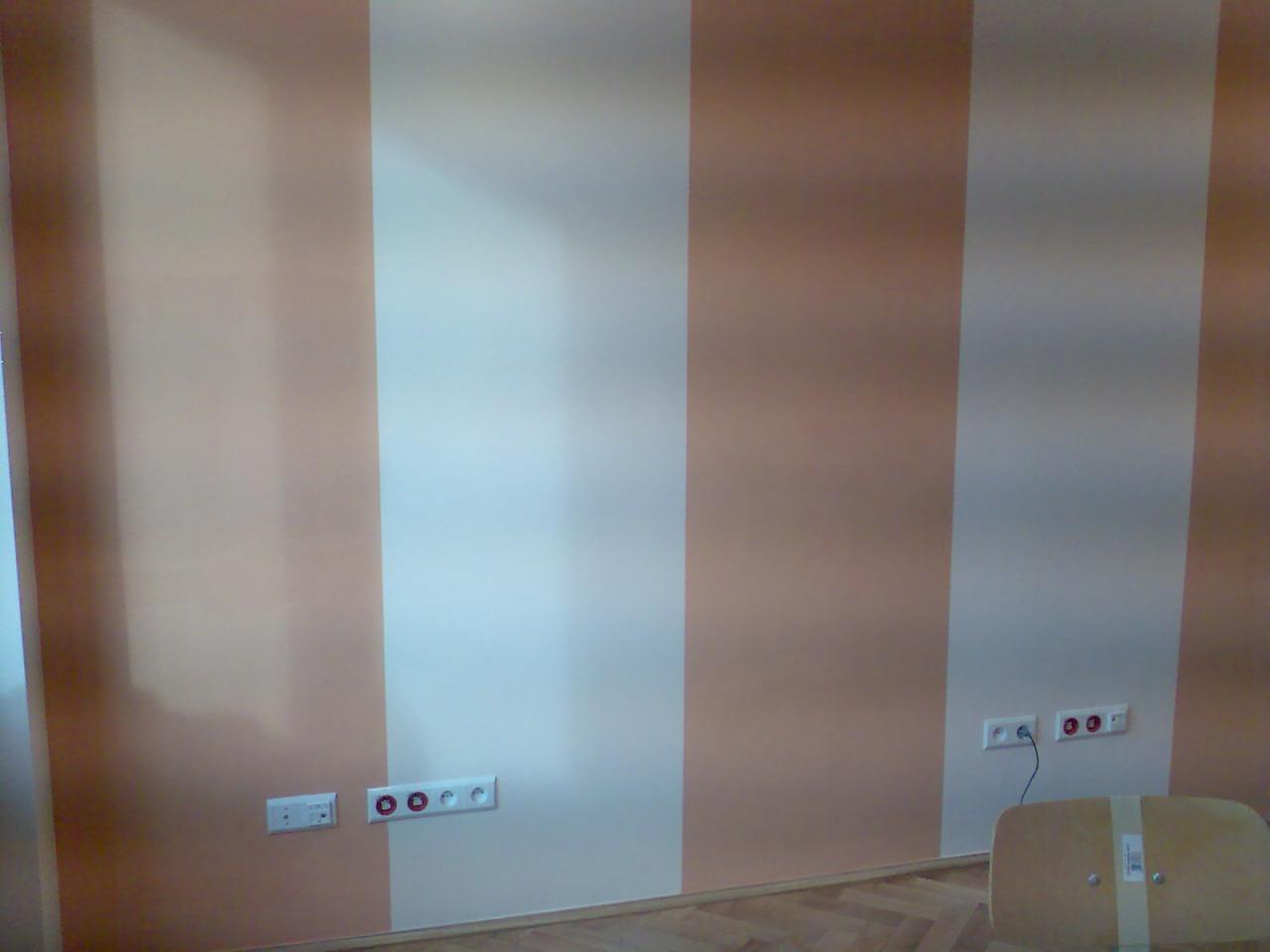 Pomalowana ściana w pionowe pasy w odcieniach beżu i bieli, z widocznymi gniazdkami elektrycznymi na dole, fragment podłogi i krzesła.