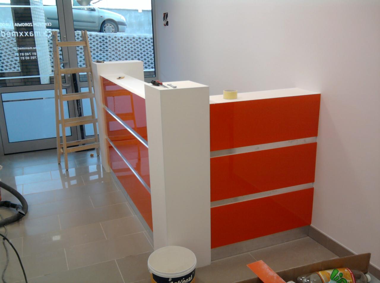 Nowoczesna recepcja z pomarańczowymi panelami i białymi elementami, w trakcie montażu, widoczna drabina, narzędzia i akcesoria malarskie, jasna podłoga z płytek.