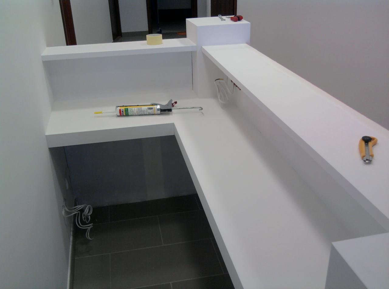 Biała zabudowa meblowa w trakcie montażu, widoczne narzędzia: tuba z klejem, nóż tapicerski, klucze, taśma malarska, wystające przewody elektryczne.